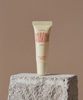 A cream-colored Suavina original lip balm tube stands upright on a textured stone block against a solid taupe background.