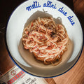 A bowl of spaghetti in a red sauce with the text "metti altri due due" on the rim, on a wooden table.
