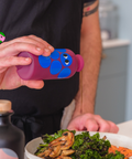 A person in a black apron drizzles a purple bottle of Yiayia and Friends Aged Balsamic Vinegar over a plate of vegetables.