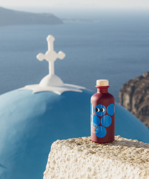 A purple bottle of Yiayia and Friends Aged Balsamic Vinegar with blue circles sits on a stone ledge in front of a blue dome and white cross overlooking the sea.
