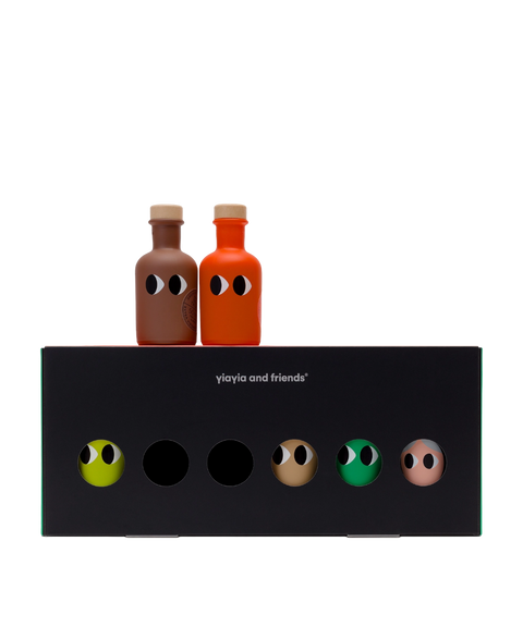 Two small bottles of olive oil with cartoon eyes, one brown and one orange, displayed on top of a black box labeled 'Yiayia and Friends' against a white background.