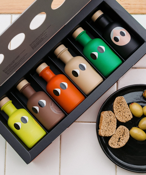 A set of six small bottles of Yiayia & Friends olive oil with cartoon eyes in a black gift box on a tiled surface next to a plate of bread and olives.