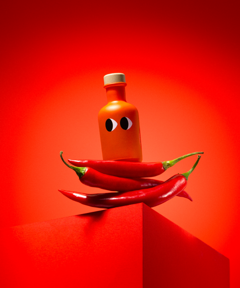A red bottle of Yiayia & Friends chili olive oil with a wood stopper balanced on chili peppers on a pedestal.