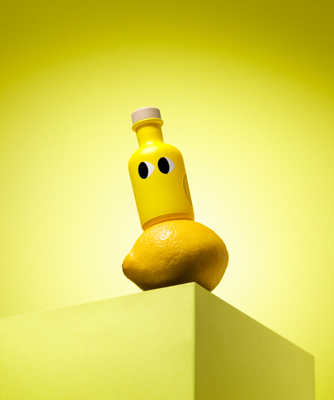 A yellow bottle of Yiayia & Friends lemon olive oil with a wood stopper balanced on a lemon on a pedestal.