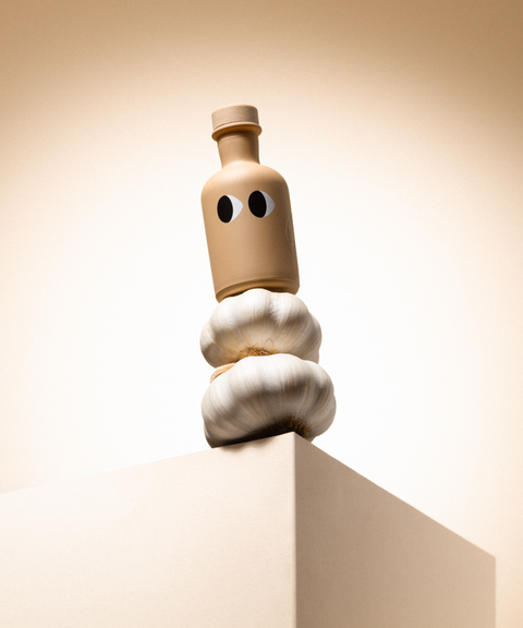 A tan bottle of Yiayia & Friends garlic olive oil with a wood stopper balanced on garlic on a pedestal.