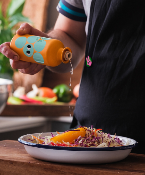 A person in a black apron holds an orange bottle of Yiayia and Friends White Balsamic Vinegar over a salad in a bowl.