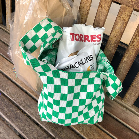 Green and white checkered tote with snacks coming out of the top on a wood bench.